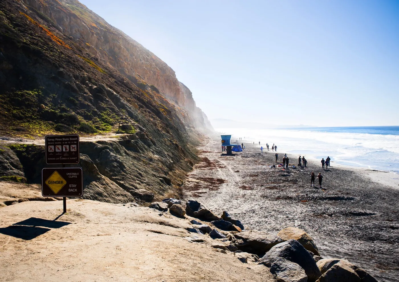 A tall sandstone cliff on the left above a wide beach, with people walking along the water's edge on a bright hazy day